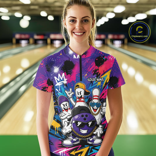 A woman stands in a bowling alley wearing a colorful graphic quarter zip shirt with cartoon bowling characters and a purple bowling ball. The background shows blurred bowling lanes and pins.