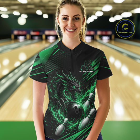 A woman stands in a bowling alley wearing a black short-sleeve bowling quarter zip shirt with a green and black dragon graphic, bowling ball and pins, and a green glow effect.