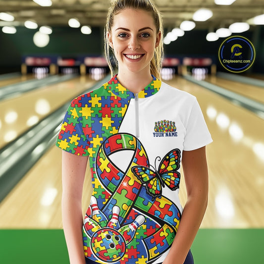 Female model in a bowling lane wearing a white quarter-zip shirt, autism ribbon and butterfly artwork with bowling ball striking pins design.