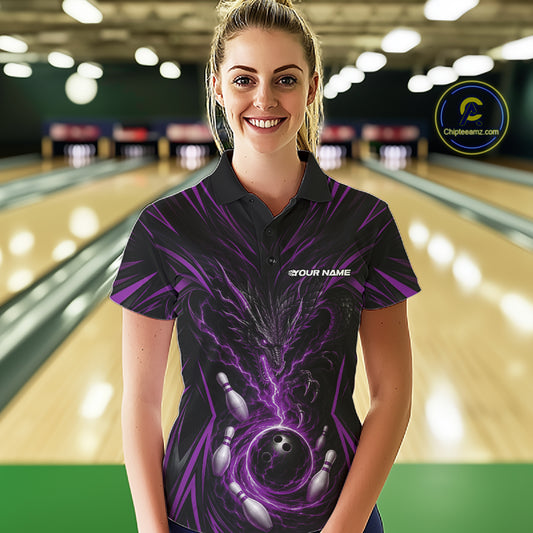 A woman stands in a bowling alley wearing a black short-sleeve polo shirt with a purple and black bowling-themed graphic print. The shirt features a large bowling ball and pins design with purple lightning effects