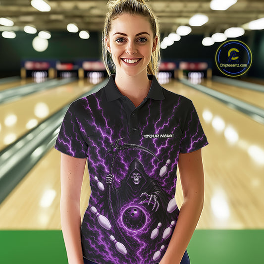 A woman stands in a bowling alley wearing a black short-sleeve polo shirt with a purple and black grim reaper graphic, featuring lightning, bowling pins, and a bowling ball.
