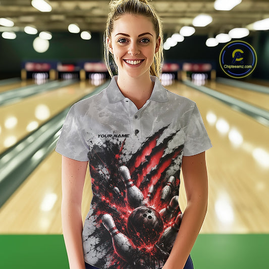 Front view of a female model wearing a gray bowling polo shirt featuring a dramatic red bowling ball and pins graphic with claw-mark artwork at a bowling lane.