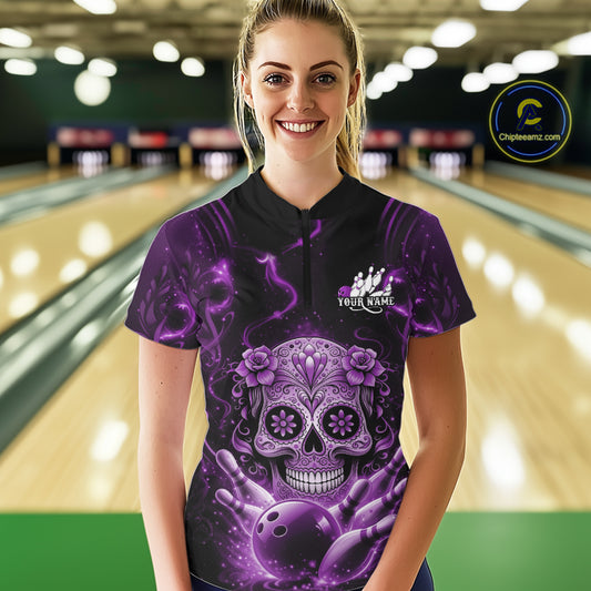 Female model at a bowling lane wearing a black and purple bowling quarter-zip shirt with floral sugar skull artwork, bowling pins, and a bowling ball motif.