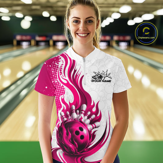 A woman stands in a bowling alley wearing a custom short-sleeve bowling shirt. The shirt is white with a vibrant magenta pink flame and bowling ball graphic on the front