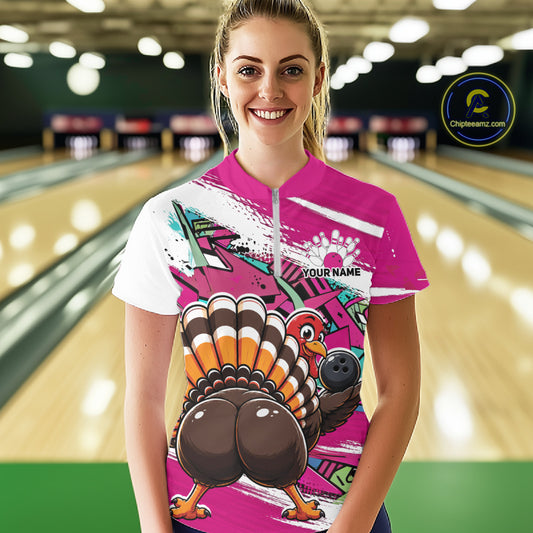 Front view of a happy female model in a bowling alley wearing a pink custom quarter zip bowling shirt with a funny graffiti turkey holding a bowling ball, paint splatter accents, and a personalized name.