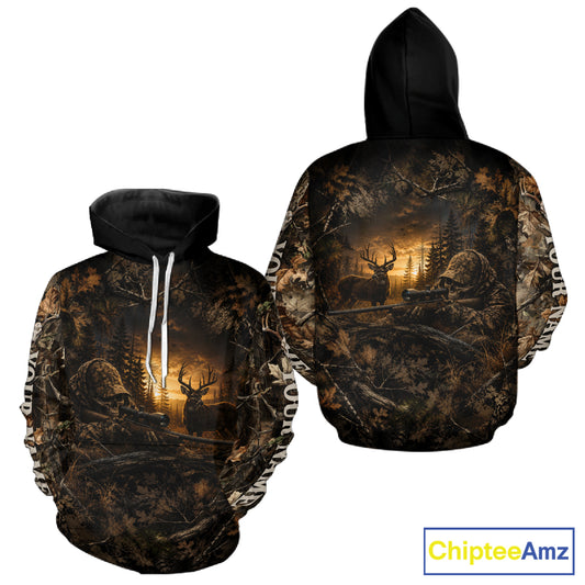 Front and back views of a black pullover hoodie featuring an all-over woodland deer hunting print with a golden sunset, deer silhouettes, and camouflage-style branches. The hoodie has white drawstrings on the front view.