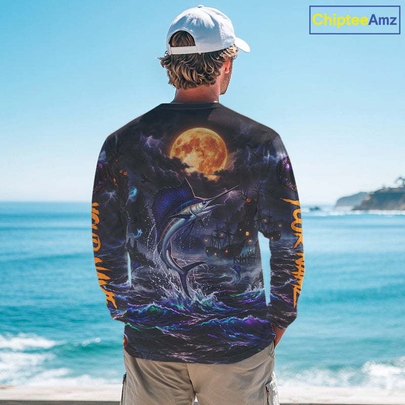 A man stands with his back to the camera wearing a long-sleeve graphic shirt featuring a dramatic print of a marlin jumping from the ocean under a large full moon, with blue and orange tones. The background shows a sunny beach scene with the ocean and sky.