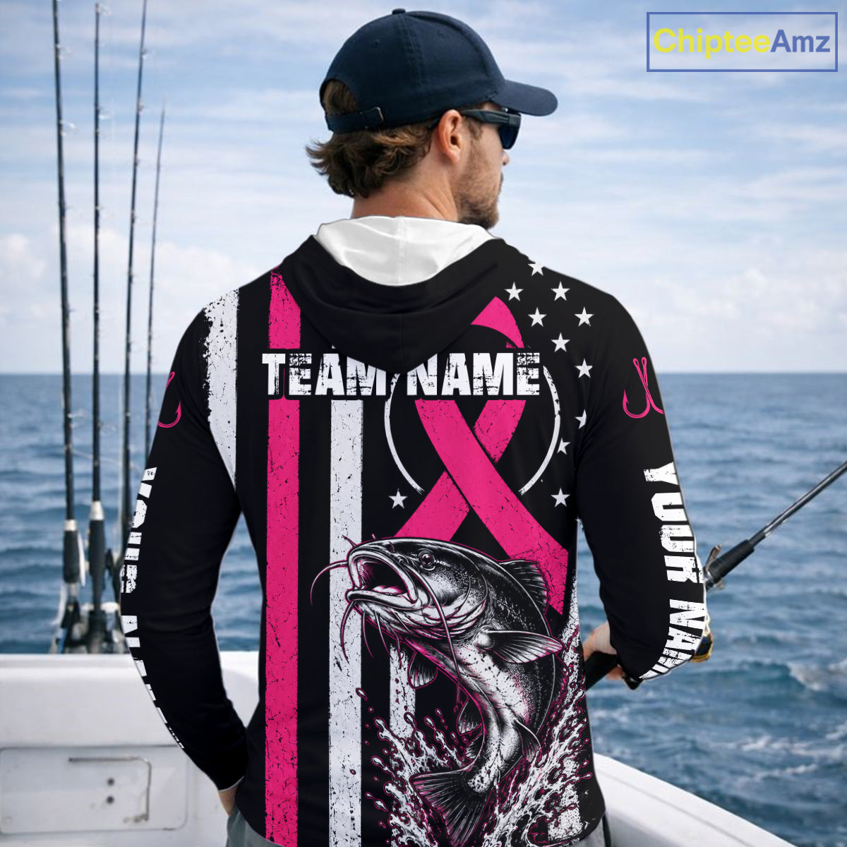 A man on a boat shown from behind wearing a custom black long-sleeve fishing hoodie with a pink and white striped design, a pink ribbon graphic, white stars, and a large fish print on the back. The hoodie includes customizable text placeholders