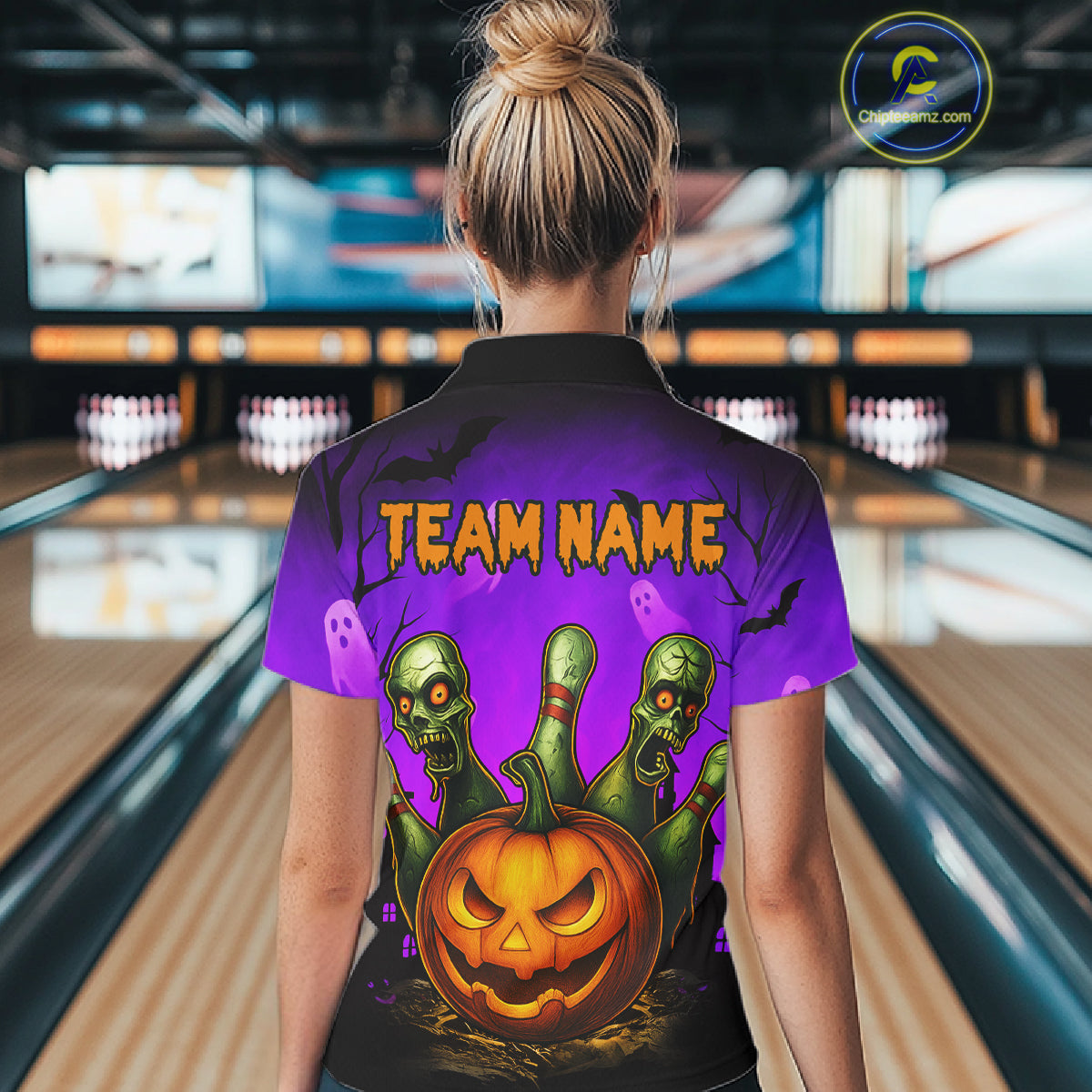 A woman viewed from behind wearing a purple short-sleeve polo shirt with a Halloween-themed graphic print on the back. The design features the text 'TEAM NAME' in orange above three green zombie heads emerging from a glowing jack-o'-lantern pumpkin.