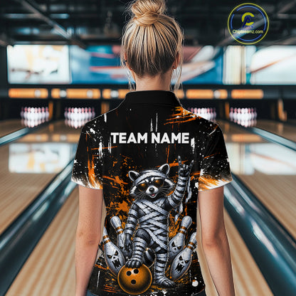 A woman viewed from behind wearing a black bowling-themed short-sleeve collared shirt with a large raccoon graphic and the text 'TEAM NAME' on the back, standing in a bowling alley.