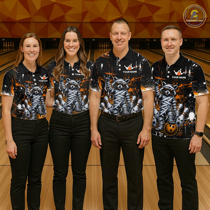 Four people standing together in a bowling alley wearing matching black short-sleeve polo shirts with a large gray raccoon and orange abstract graphic print, paired with black pants. The shirts have white placeholder text and a circular logo in the top right corner.