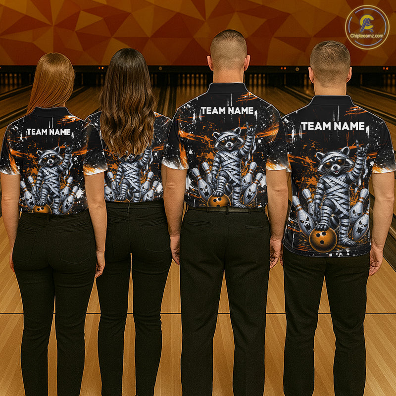 Four people standing with their backs to the camera in a bowling alley, wearing matching black short-sleeve bowling shirts with a detailed graphic of a mummy wrapped in bandages, skulls, and bowling pins, with 'TEAM NAME' printed on the back.