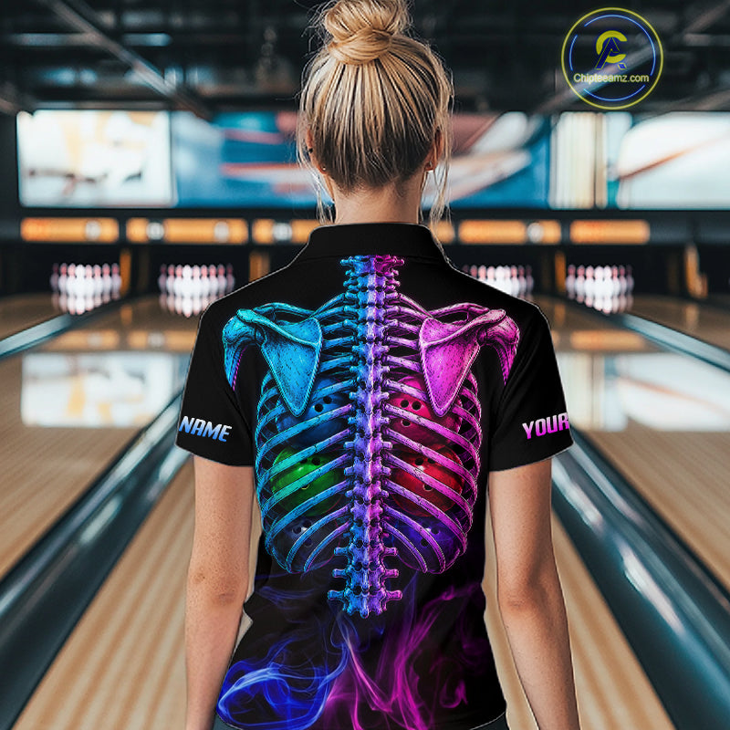 A woman viewed from behind wearing a black short-sleeve polo shirt with a large colorful skeletal ribcage graphic on the back, standing in a bowling alley. 