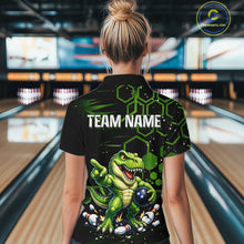 Load image into Gallery viewer, A woman viewed from behind wearing a black short-sleeve bowling shirt with a green cartoon dinosaur graphic and the text 'TEAM NAME' on the back, standing in a bowling alley.
