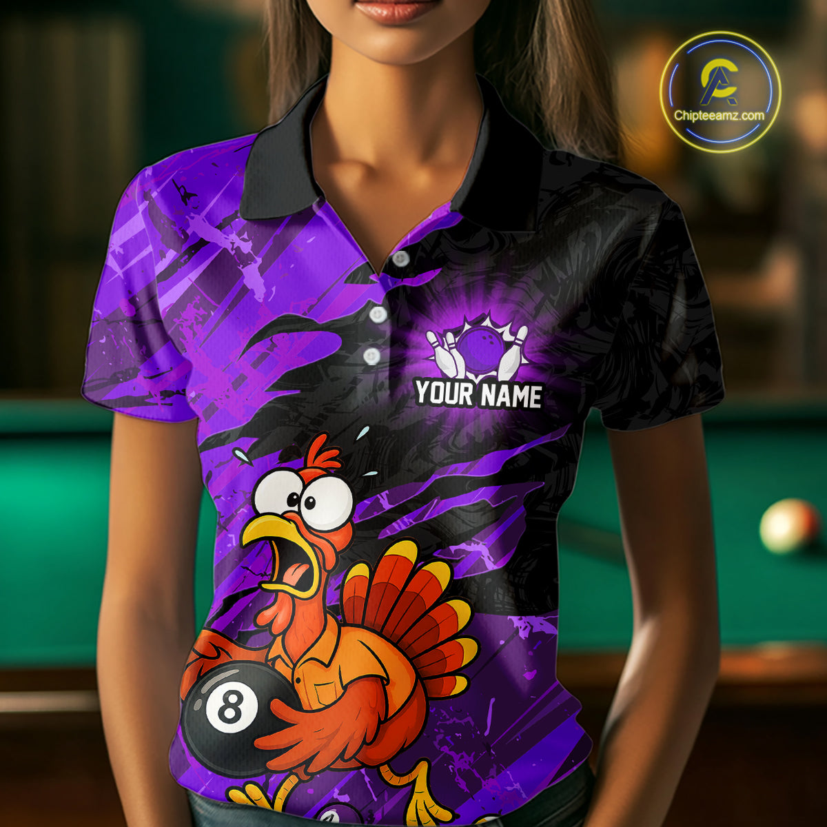 A woman wearing a short-sleeve polo shirt with a vibrant purple and black graphic design featuring a cartoon turkey holding an 8-ball and the text 'YOUR NAME' and bowling pins. The background shows a blurred pool table.