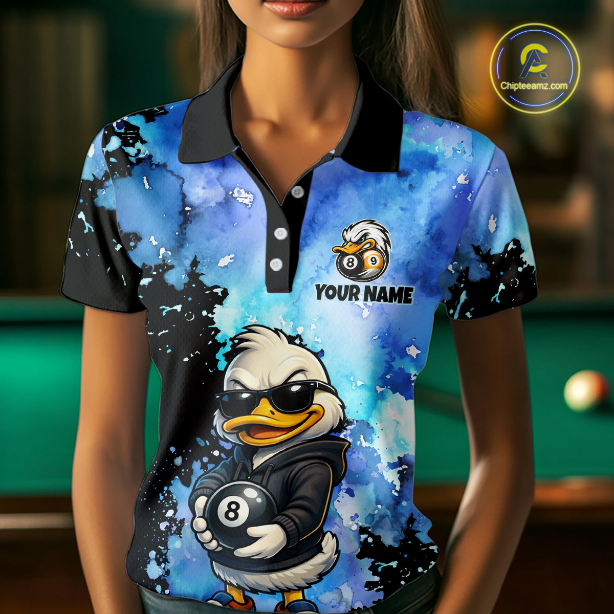 A woman wearing a short-sleeve polo shirt with a blue and black watercolor-style graphic print featuring a cartoon duck wearing sunglasses and holding an 8-ball, with the text 'YOUR NAME' and a logo on the chest. The background is a blurred indoor pool room setting.