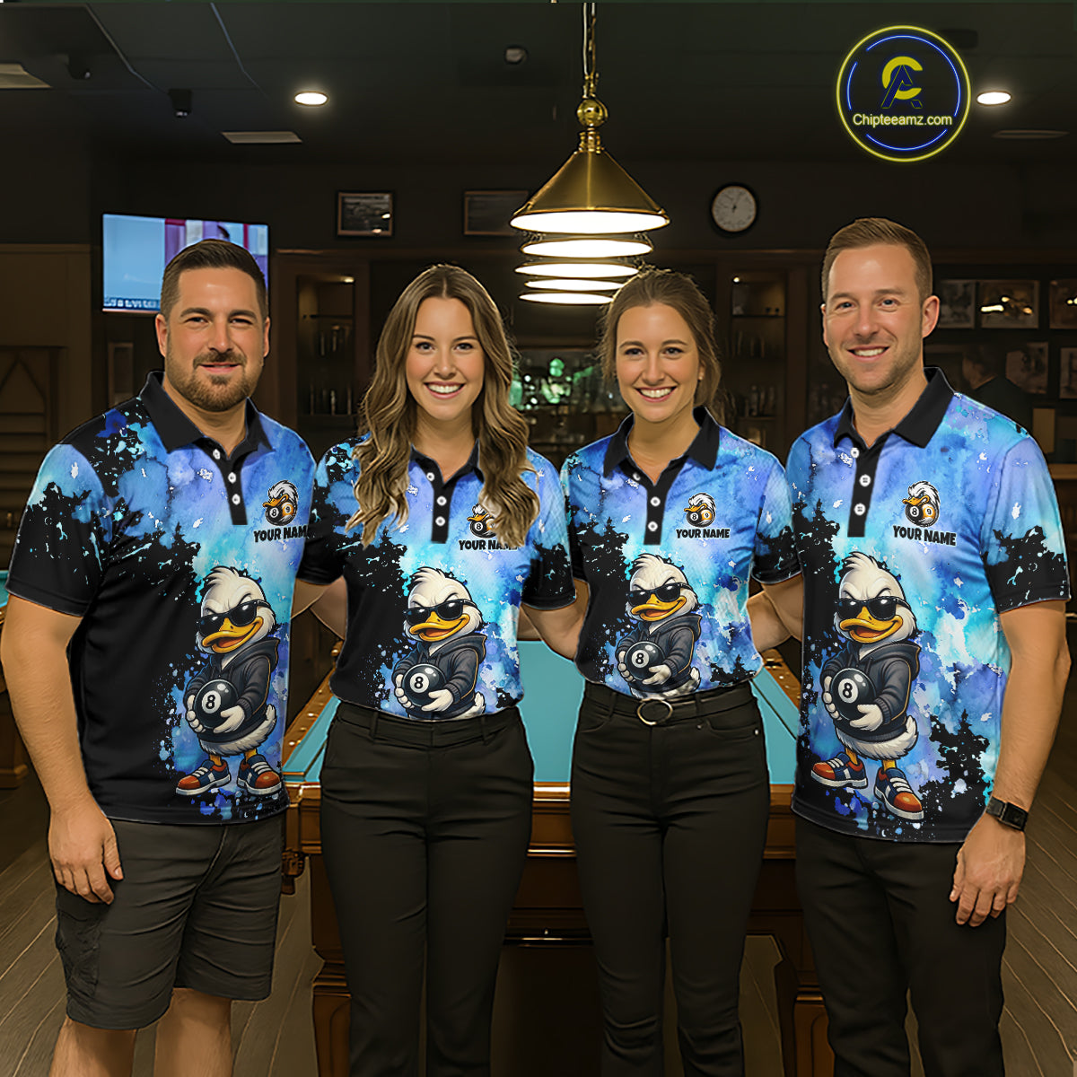 Four people standing together in a dimly lit indoor venue, wearing matching blue and black polo shirts with a large cartoon duck graphic and the text 'YOUR NAME'  on the chest.