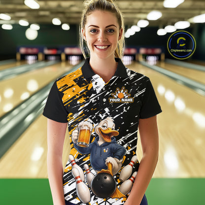 A woman stands in a bowling alley wearing a black short-sleeve polo shirt with a colorful cartoon duck graphic holding a bowling ball and beer, plus a 'YOUR NAME' customization area on the chest. Bowling lanes are blurred in the background