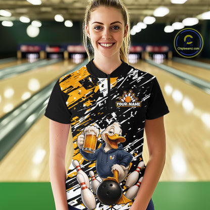A woman stands in a bowling alley wearing a custom-designed bowling-themed polo shirt. The shirt features a black base with yellow and white brushstroke patterns, a cartoon duck character holding a beer and bowling pins, a black bowling ball, and a customizable name patch.