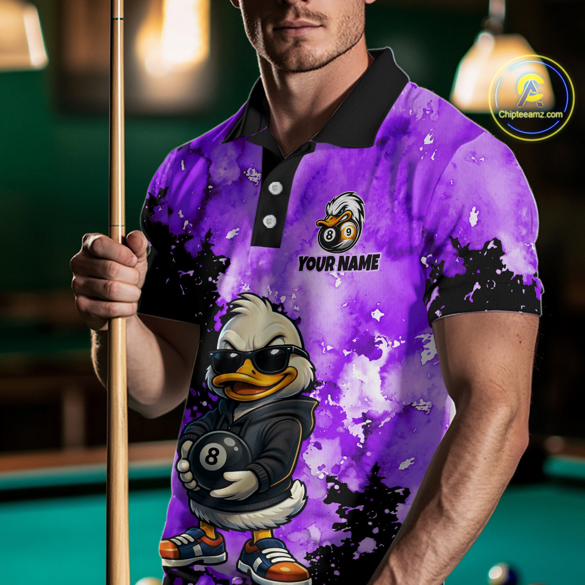 A man wearing a custom purple and black watercolor-patterned polo shirt with a cartoon duck graphic holding an 8-ball and the text 'YOUR NAME'. He is holding a pool cue in a dimly lit pool hall environment.