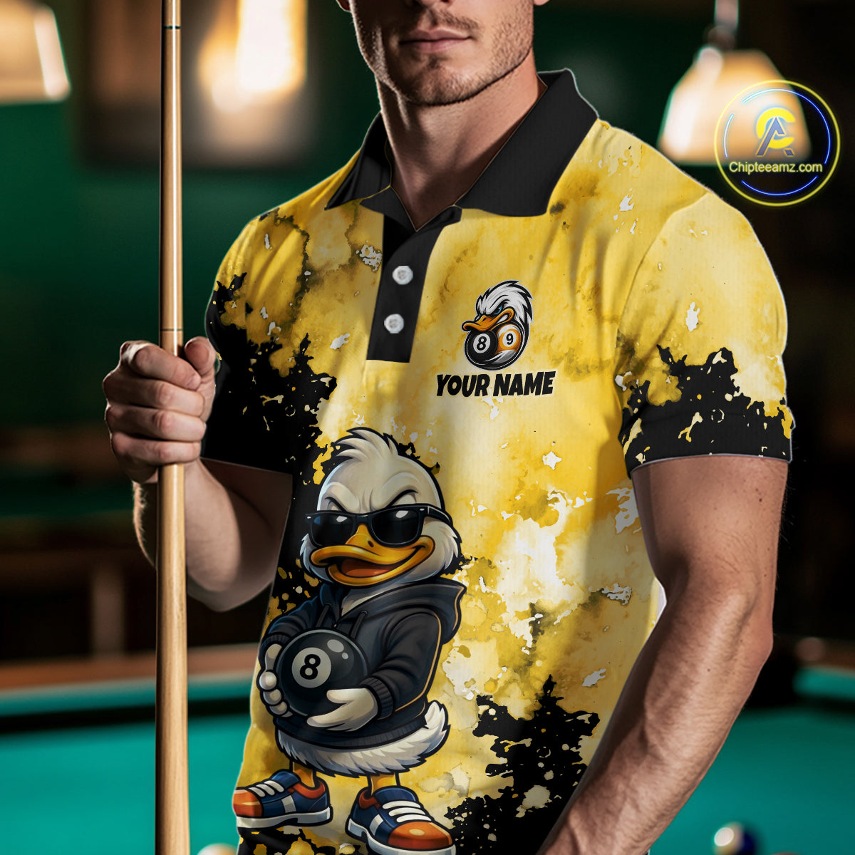 A man wearing a yellow and black polo shirt with a cartoon duck playing billiards graphic, holding a pool cue in a dimly lit pool hall. 
