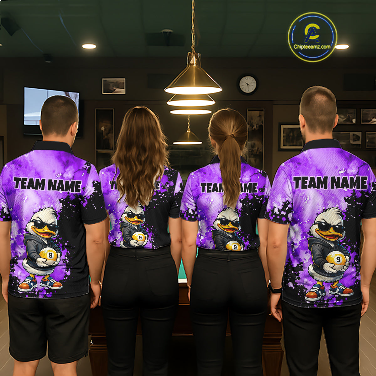 Four people standing with their backs to the camera in a dimly lit indoor room, wearing matching purple and black short-sleeve shirts with a cartoon duck graphic and the text 'TEAM NAME' across the upper back. 