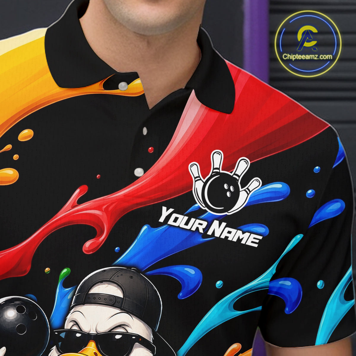 A close-up of a person wearing a black short-sleeve polo shirt with a colorful splash design in red, blue, yellow, and orange, plus a bowling pin and duck graphic. The shirt has a white 'YOUR NAME' placeholder print on the chest 