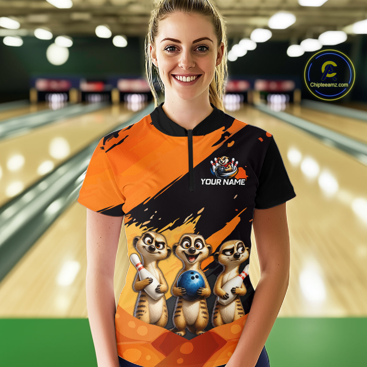 A woman stands in a bowling alley wearing a custom short-sleeve bowling jersey. The jersey features a black and orange paint-splatter design with a cartoon meerkat graphic holding a bowling ball on the front, and the text 'YOUR NAME' on the chest.