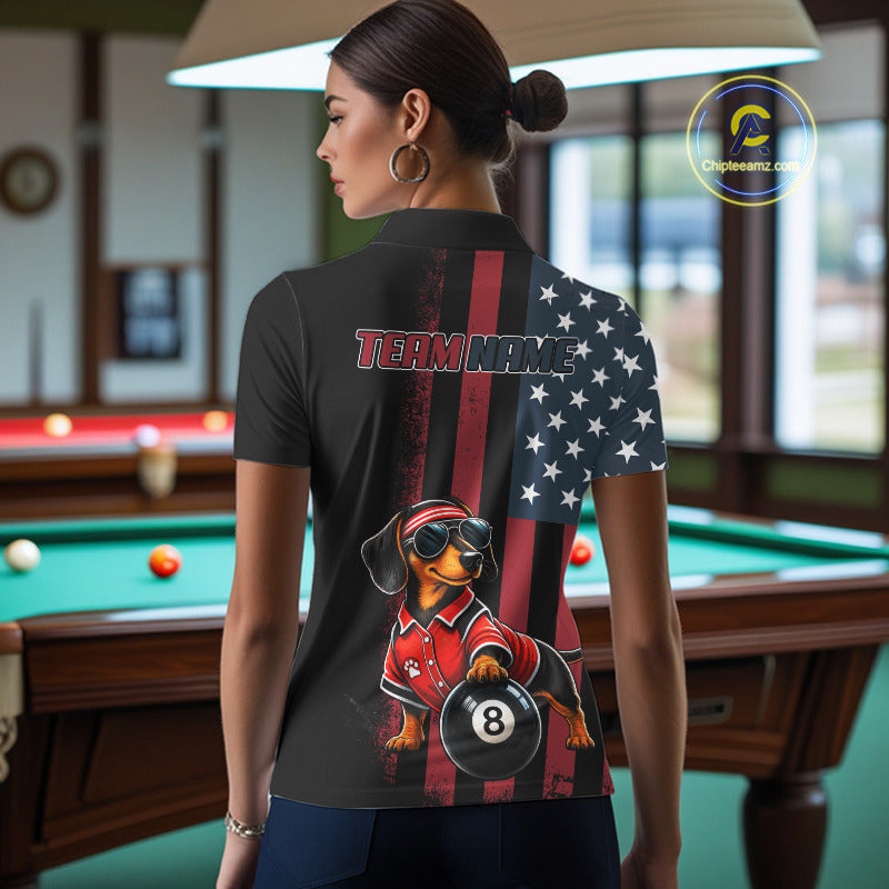 A woman stands in a billiards room wearing a black short-sleeve polo shirt with a custom American flag and dachshund graphic on the back. The shirt features a vertical split design with a red and black American flag on one side and a white star field on the other, with a dachshund wearing a red polo and sunglasses holding an 8-ball.