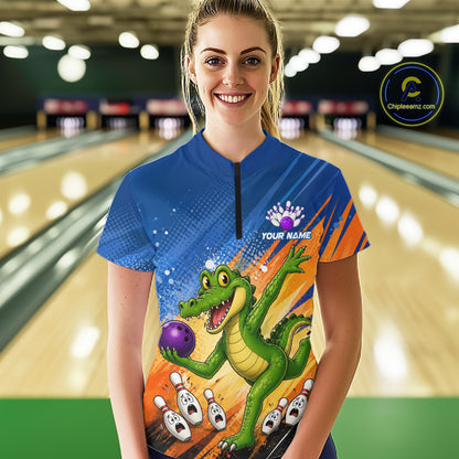 A woman stands in a bowling alley wearing a short-sleeve bowling jersey with a colorful cartoon alligator holding a purple bowling ball, orange and blue graphic panels, and a black zipper at the neck. 