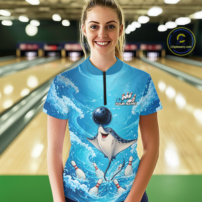 A woman stands in a bowling alley wearing a bright blue short-sleeve bowling jersey with a printed graphic of a manta ray catching a bowling ball, bowling pins, and waves. The jersey has a black zipper at the collar and a customizable name patch on the chest.