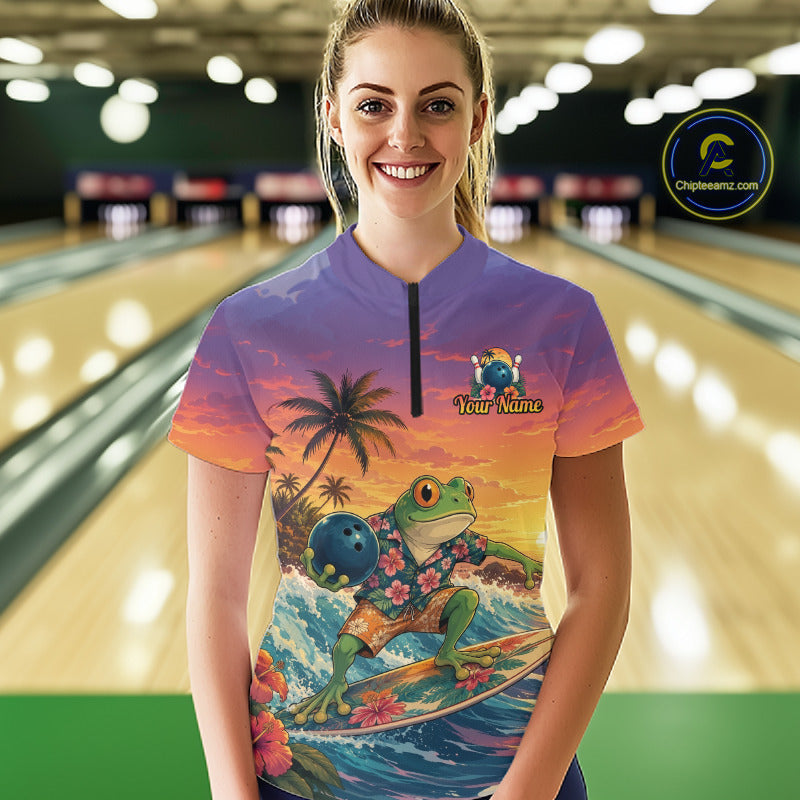 A woman stands in a bowling alley wearing a short-sleeve quarter-zip polo shirt with a colorful tropical frog surfing graphic. The shirt features a purple-to-orange sunset gradient, palm trees, flowers, and a green frog holding a bowling ball.