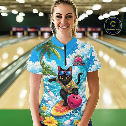 A woman stands in a bowling alley wearing a short-sleeve collared quarter-zip shirt with a colorful all-over print featuring a black cat surfing on a yellow board with a pink bowling ball, palm trees, clouds, and bowling pins. The background shows blurred bowling lanes.