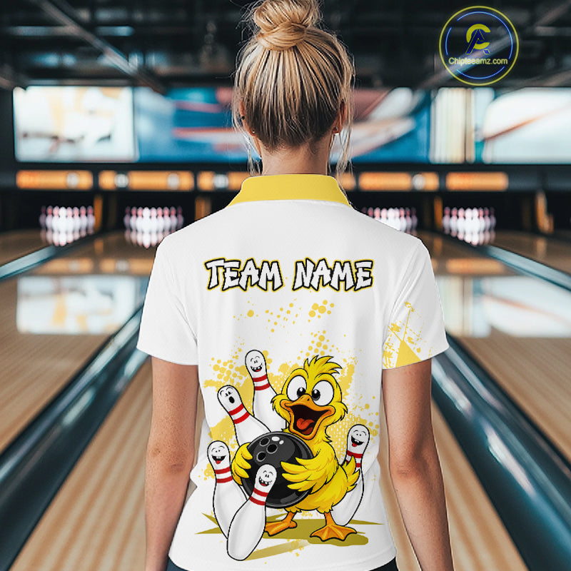 A woman viewed from behind wearing a white bowling-themed t-shirt with a yellow collar, featuring a cartoon yellow duck holding a bowling ball and pins, and the text 'TEAM NAME' on the back. The background shows a blurred bowling alley with lanes and pins.