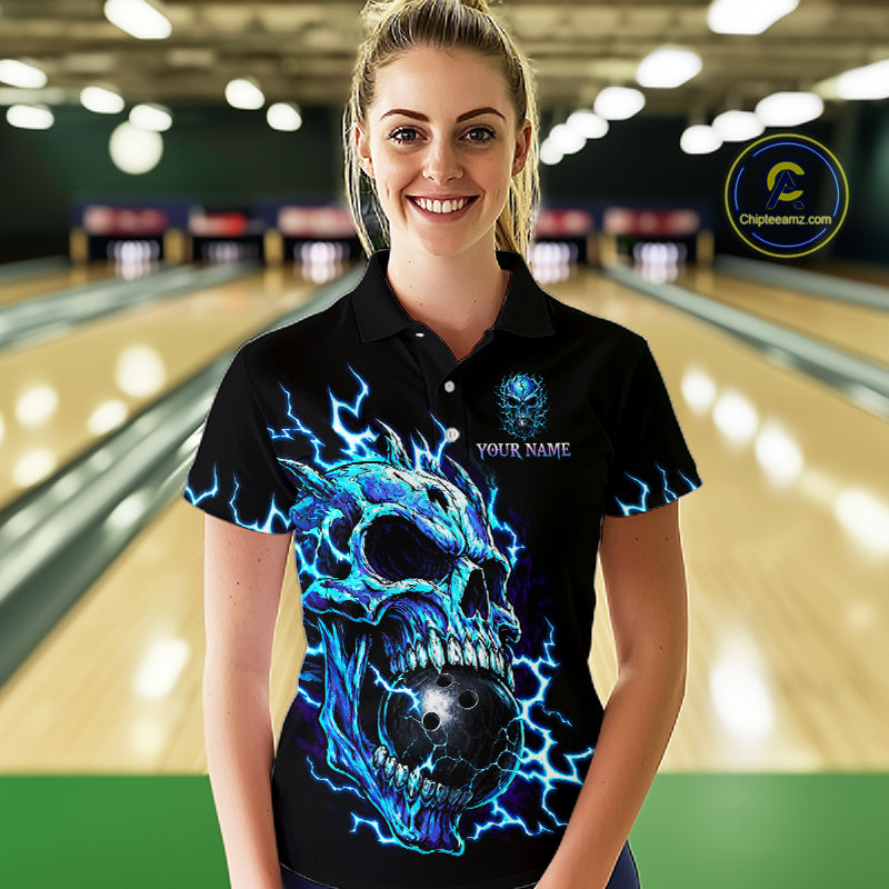 A woman stands in a bowling alley wearing a black short-sleeve polo shirt with a large blue and black skull graphic and lightning effects. The shirt has a small crest on the left chest and 'YOUR NAME' text on the right chest.