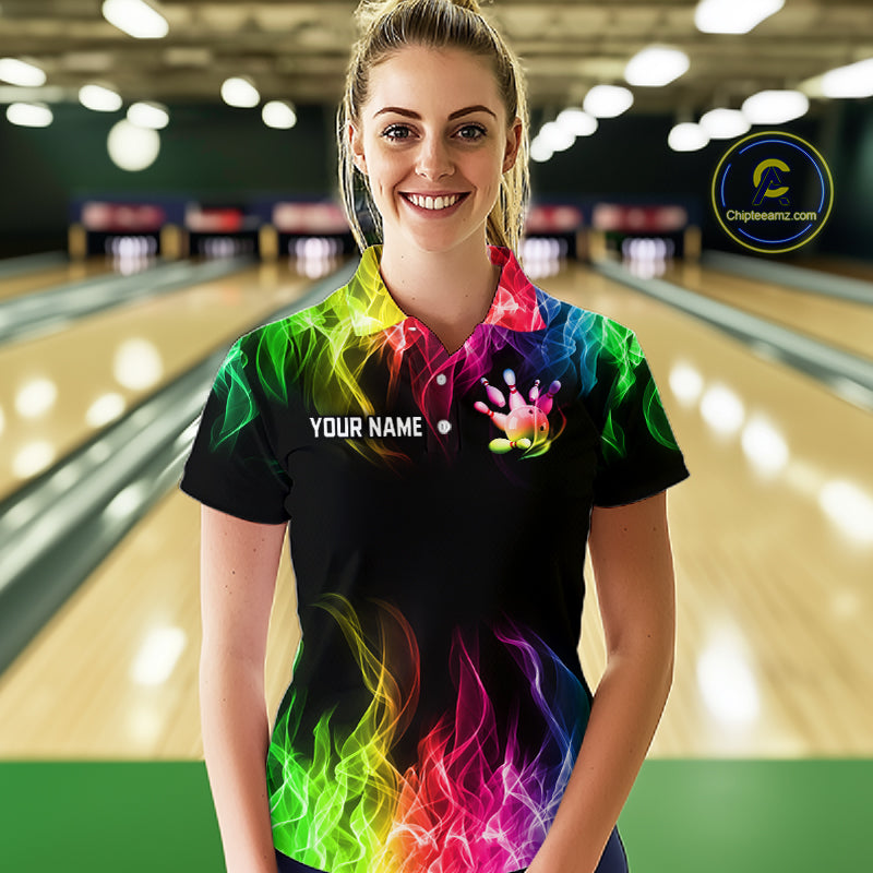 A woman stands in a bowling alley wearing a black short-sleeve polo shirt with a vibrant neon rainbow flame and handprint graphic. The shirt features a customizable name print on the chest.