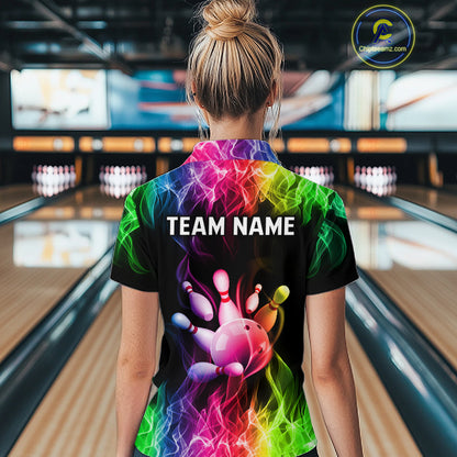 A woman viewed from behind wearing a black short-sleeve bowling shirt with a vibrant rainbow graphic of a bowling ball and pins, with 'TEAM NAME' printed across the back. The background shows blurred bowling alley lanes.