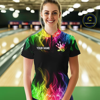 A woman stands in a bowling alley wearing a black short-sleeve bowling shirt with a vibrant rainbow flame pattern and customizable text on the chest. 