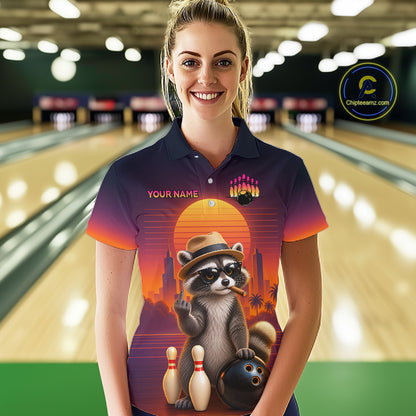 A woman wearing a custom bowling-themed short-sleeve polo shirt with a colorful raccoon graphic, standing in a bowling alley. The shirt features a sunset design with a raccoon wearing a hat and holding a bowling ball next to bowling pins.
