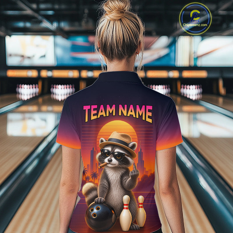 A woman viewed from behind wearing a bowling-themed short-sleeve polo shirt in a bowling alley. The shirt features a colorful graphic of a raccoon wearing a hat and sunglasses, holding a bowling ball next to bowling pins, with the text 'TEAM NAME' printed across the upper back.