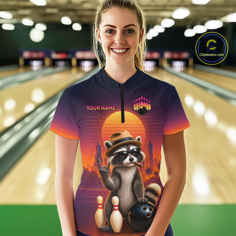A woman wearing a custom short-sleeve bowling-themed polo shirt with a raccoon graphic, standing in a bowling alley. The shirt features a sunset gradient background with a raccoon wearing a hat and holding a bowling ball, with 'YOUR NAME' printed on the chest.