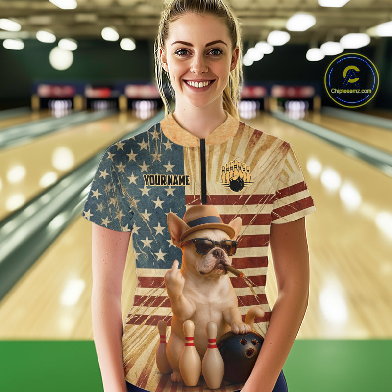 A woman stands in a bowling alley wearing a custom short-sleeve bowling jersey. The jersey features a split design: one side has a blue and beige American flag pattern with 'YOUR NAME' printed on it, and the other side shows a vintage-style American flag background with a graphic of a French Bulldog bowling pinning down bowling pins and a bowling ball.