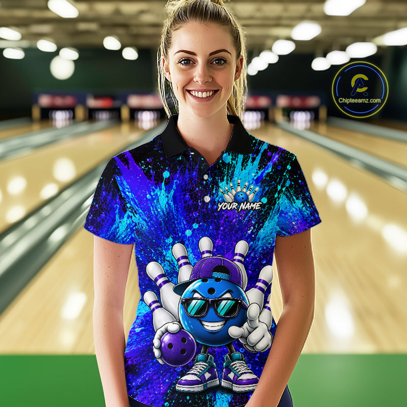 A woman stands in a bowling alley wearing a short-sleeve polo shirt with a vibrant blue and purple cosmic paint splatter design and a cartoon bowling character graphic on the front. The shirt has a black collar and the text 'YOUR NAME' printed on the chest.