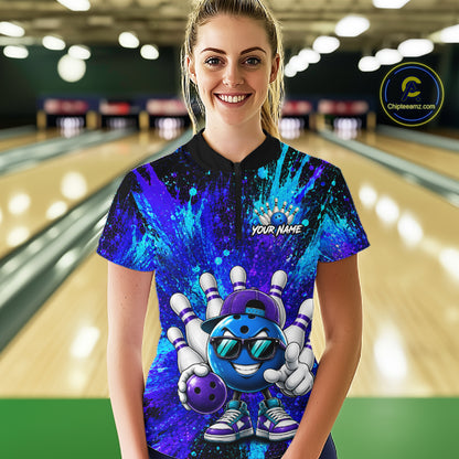 A woman stands in a bowling alley wearing a custom short-sleeve bowling t-shirt with a vibrant blue and purple cosmic paint splatter design, featuring a cartoon bowling pin and ball character on the front and a customizable name print on the chest.