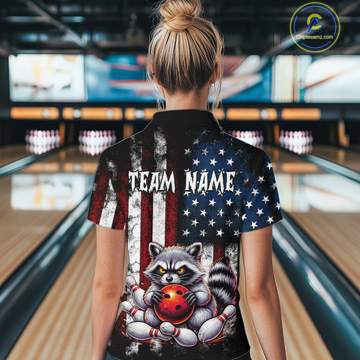 A woman viewed from behind wearing a short-sleeve bowling-themed polo shirt in a bowling alley. The shirt features a distressed American flag design with a raccoon holding a bowling ball on the back, plus the text 'TEAM NAME' .