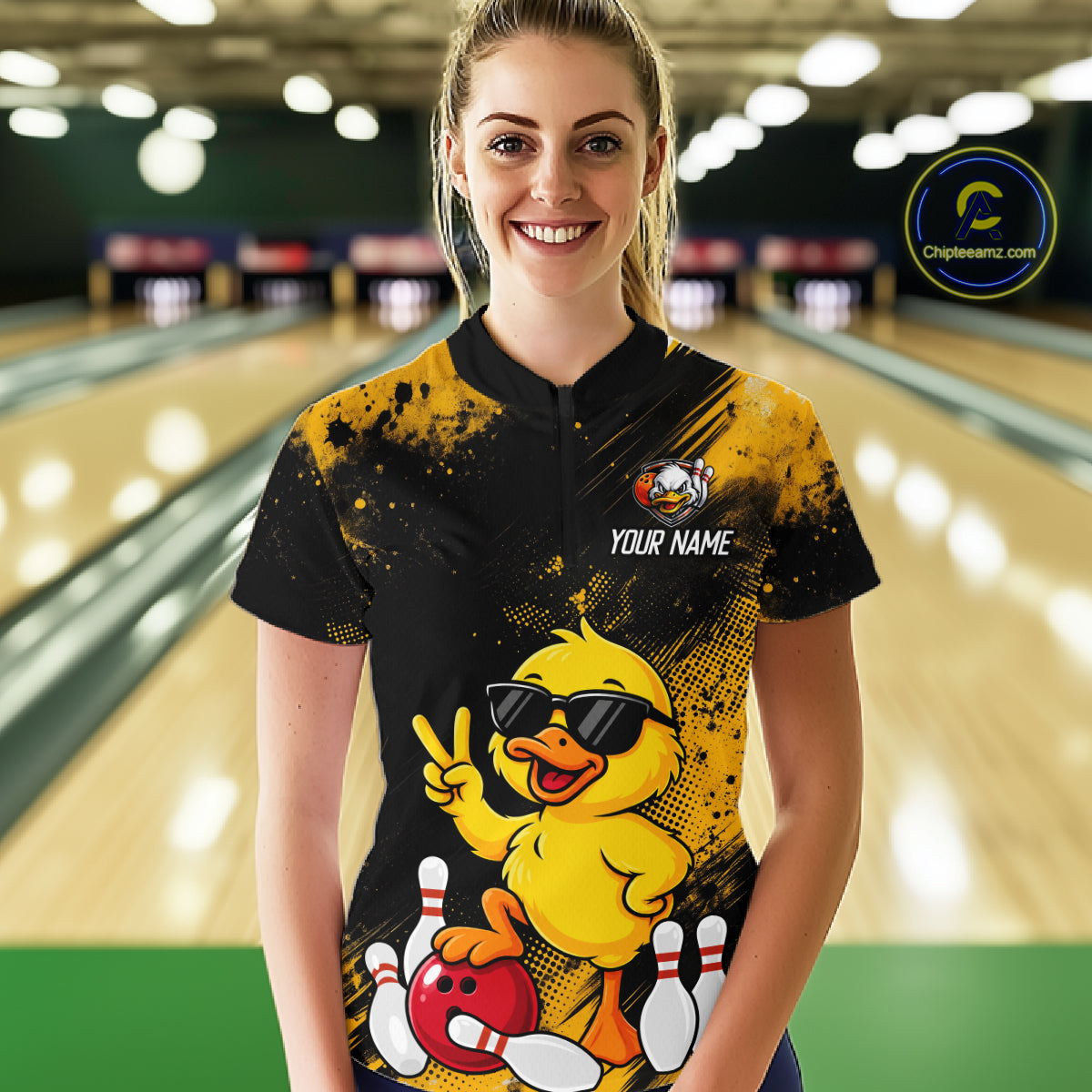 A woman standing in a bowling alley wearing a black and yellow bowling-themed graphic t-shirt with a cartoon duck character and the text 'YOUR NAME'.
