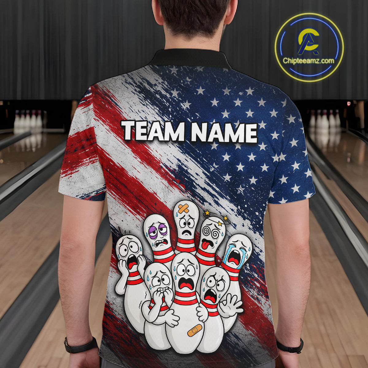 A person standing in a bowling alley wearing a short-sleeve collared shirt from the back. The shirt features a distressed American flag design in red, white, and blue, with the text 'TEAM NAME' across the upper back and a cartoon group of crying bowling pins near the lower back.