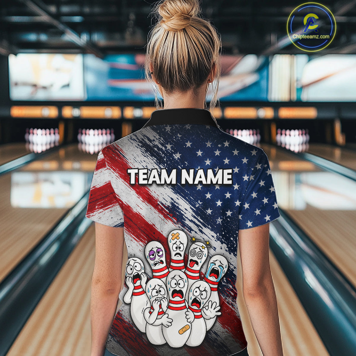A woman viewed from behind wearing a short-sleeve bowling-themed polo shirt with a patriotic red, white, and blue design, cartoon bowling pins, and the text 'TEAM NAME' on the back. The background shows a blurred bowling alley interior.