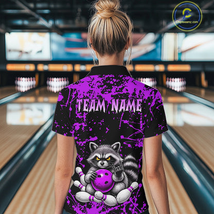 A woman viewed from behind wearing a custom bowling-themed polo shirt with a black and purple splatter design, a raccoon holding a purple bowling ball graphic, and 'TEAM NAME' printed on the back. The background shows a blurred bowling alley with lanes and pins.