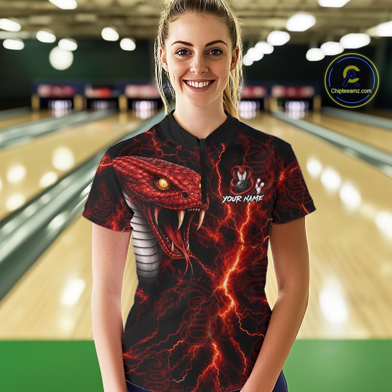 A woman stands in a bowling alley wearing a black short-sleeve polo shirt with a vivid red and black snake graphic, lightning effects, and a small bunny logo with the text 'YOUR NAME'. Bowling lanes are blurred in the background